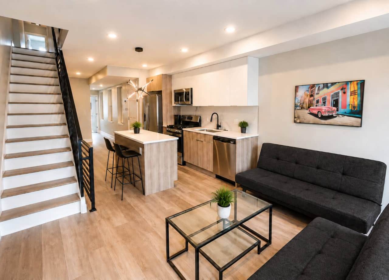 living room and kitchen featuring two black linen futon sofas, a glass coffee table, and a sleek kitchen with stainless-steel appliances, a large island, and two black barstools. The space offers plenty of lighting, stylish finishes, and comfort.