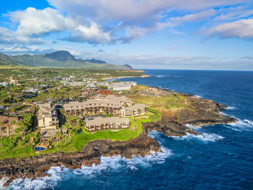 Sweeping aerial of Poipu coastline with oceanfront resort, lava rock shoreline, and lush mountain ba