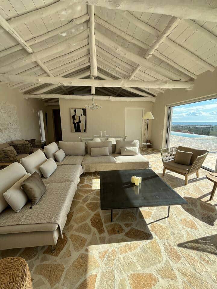 The Poolside Apartment : 
A spacious living room bathed in natural light, beneath a white-washed beamed ceiling with Mediterranean charm. Generous sofas invite relaxation, while the sea horizon unfolds beyond the openings in serene luxury.