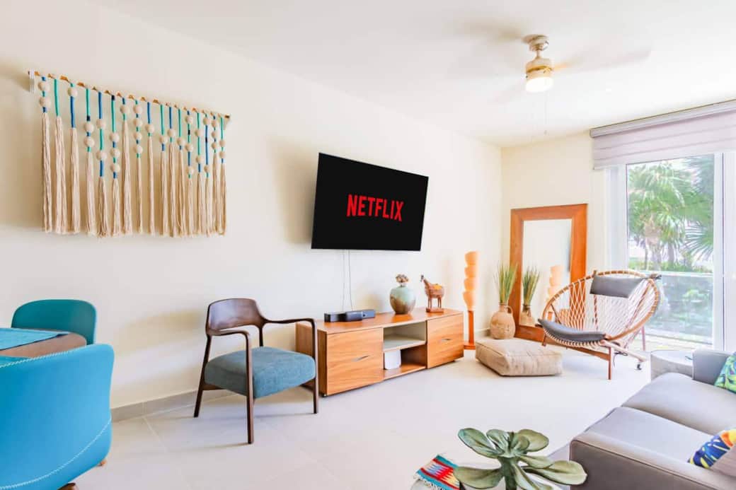 Modern living room with a Netflix-ready smart TV, comfortable seating, and large windows overlooking lush greenery, plus direct access to the private terrace to enjoy ocean views. A relaxing space to unwind and enjoy your stay.
