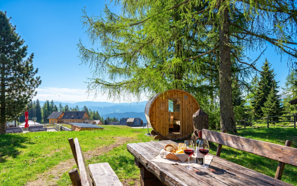 Jause mit Rotwein am Holztisch vor der Fasssauna mit Panoramablick auf die Berglodge Hochrindl