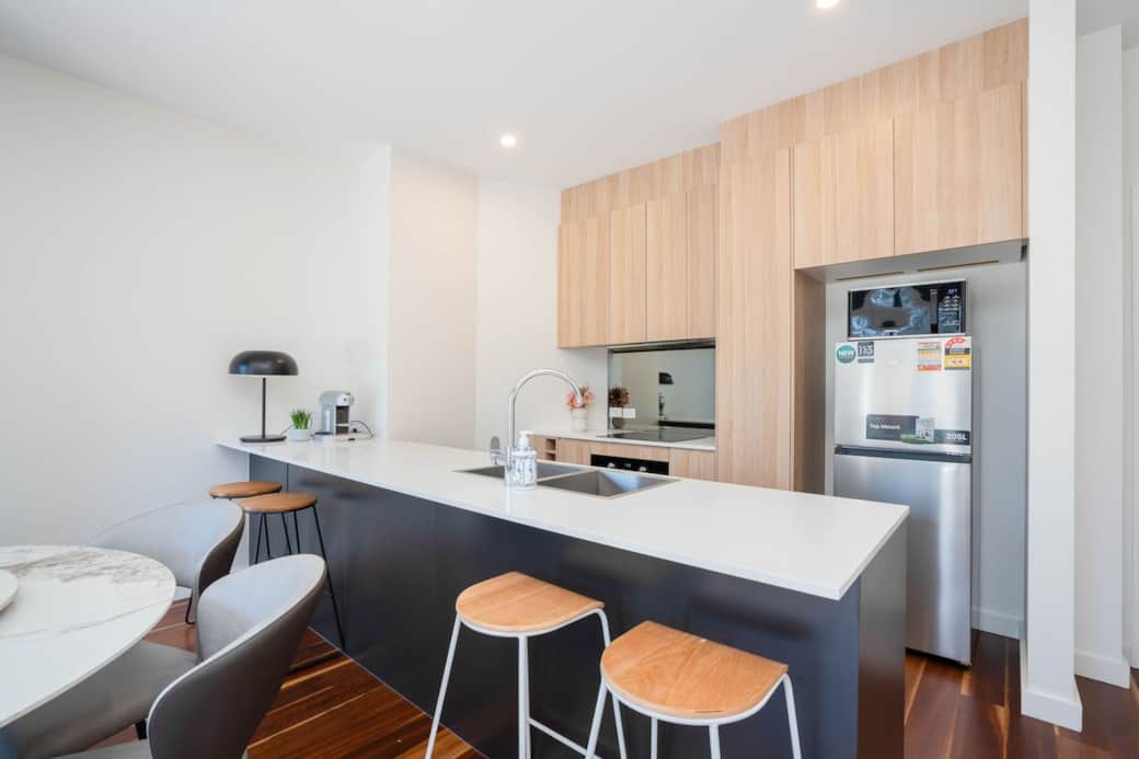 Sleek kitchen with a white countertop, wooden cabinetry, and stainless steel appliances. Includes a breakfast bar with stylish stools, perfect for casual dining. Bright and modern, the space is equipped with a sink, & espresso machine. 