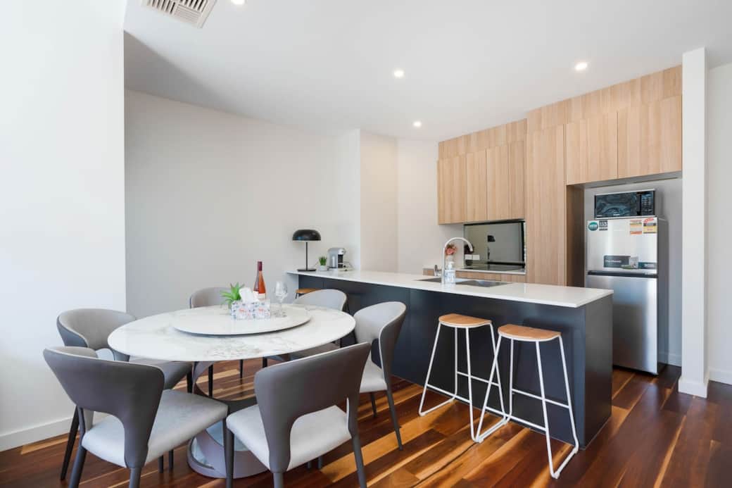 Featuring a round marble dining table with comfortable grey chairs, the kitchen has sleek wooden cabinetry, a white countertop, and bar stools for casual seating. Wooden flooring ties the space together, offering a stylish, cozy vibe.