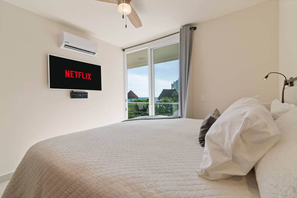 Bedroom 1 features a cozy king-size bed with soft pillows and sheets, a 55-inch Smart TV, air conditioning, a ceiling fan, and access to a private balcony with stunning ocean views.