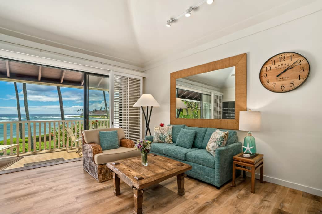 Cozy oceanfront living—sofa & rattan chair, big mirror, breezy lanai, waves and palms just beyond.