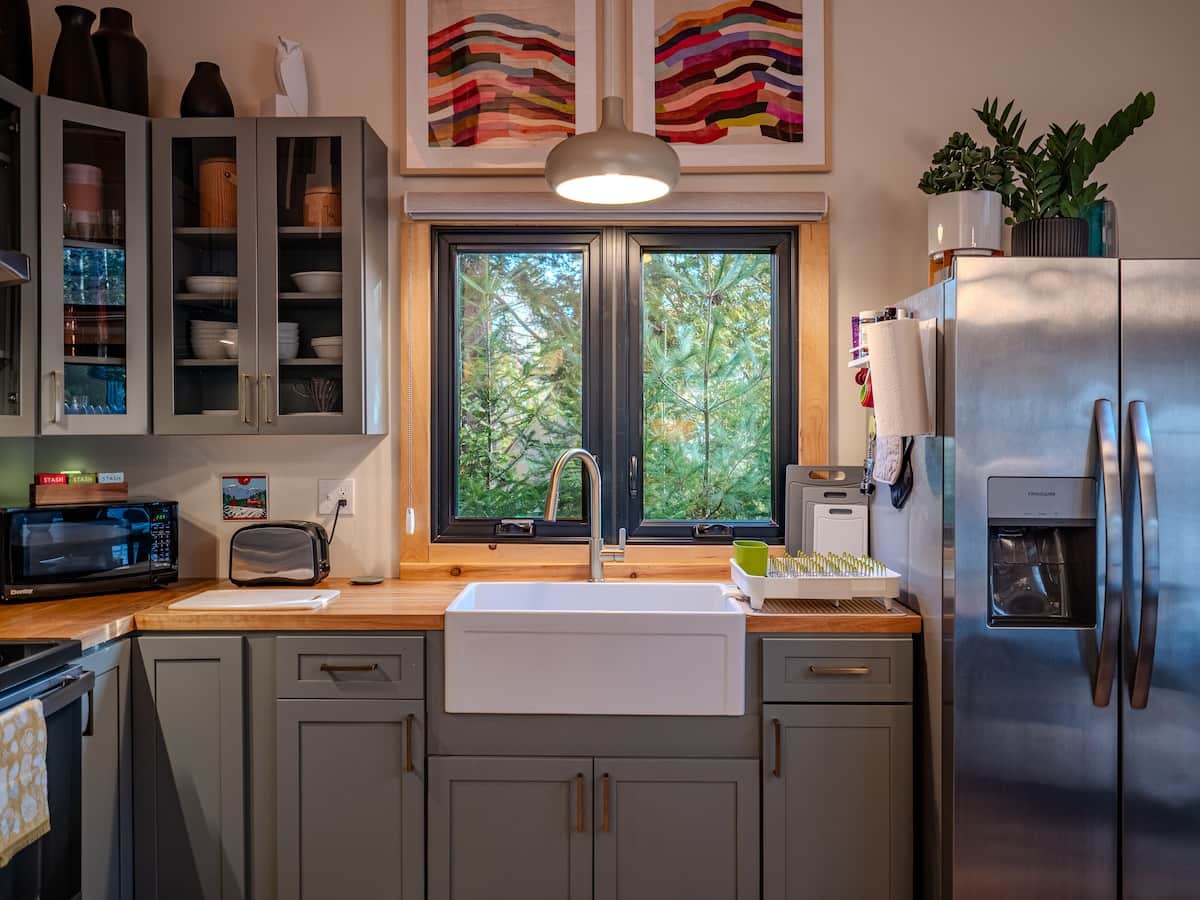 This modern rustic kitchen features glass-front cabinetry, butcher block countertops, and a large white farmhouse sink. A black-framed window offers a forest view, complemented by stainless steel appliances and colorful abstract art.