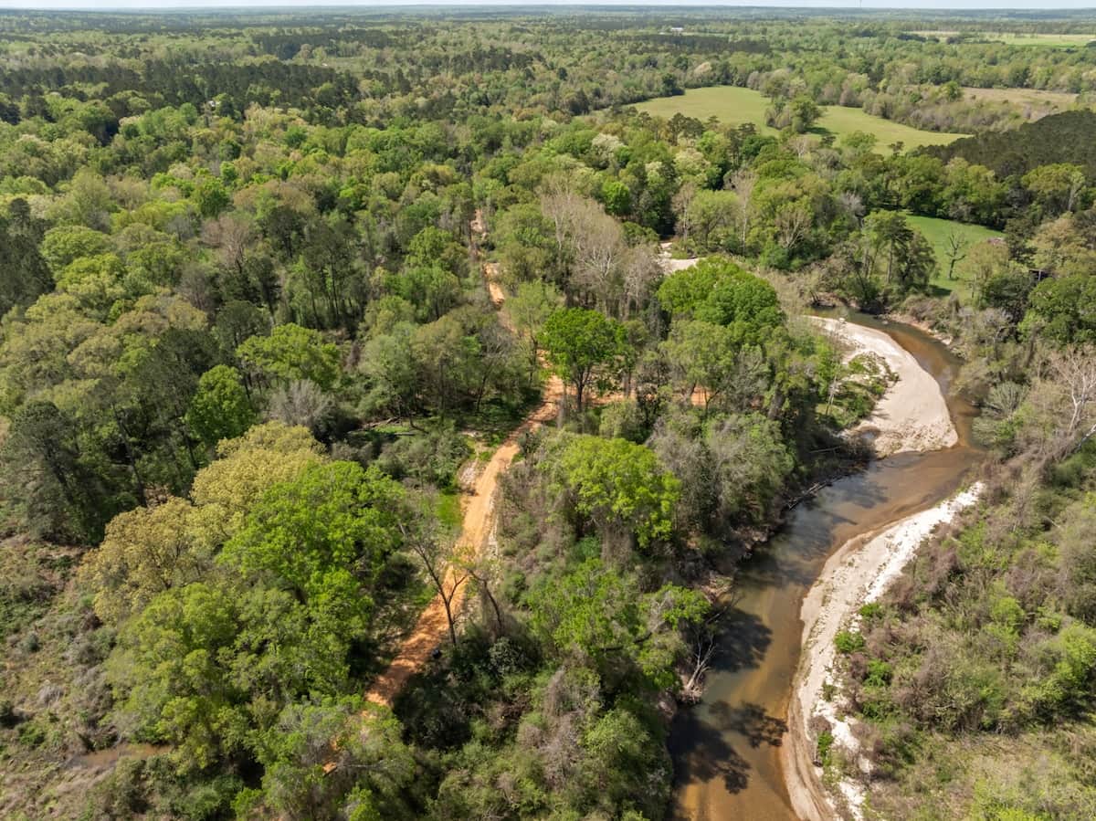 A sweeping aerial view of the property, where the creek curves through sandy banks and private sandbars below, and the red dirt road winds its way from the gate to the cabin tucked deep in the trees.
