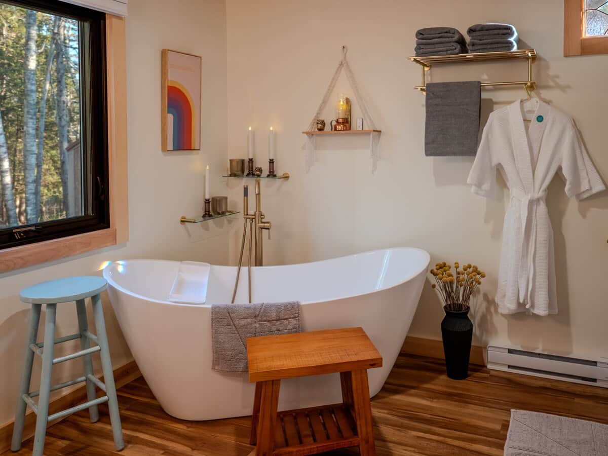 This cozy, spa-style bathroom features a white freestanding soaking tub with gold fixtures. Warm wood floors, eclectic wall art, a robe, and views of a wooded landscape create a peaceful, modern rustic retreat.