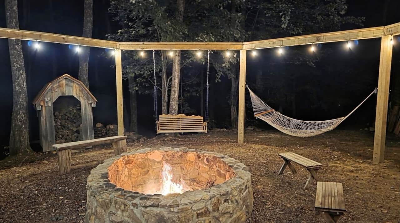 Stone firepit surrounded by a hammock stand. A wood shed in the background and string light decor.