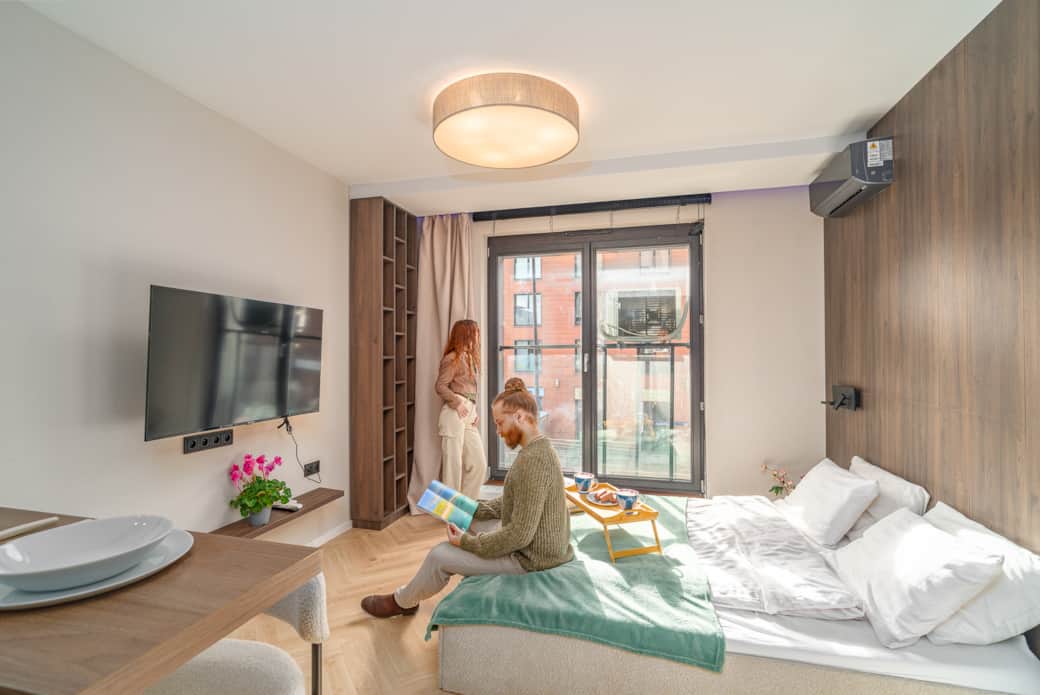 Jasna część sypialniana z dużym łóżkiem, szerokim oknem i drewnianą zabudową.