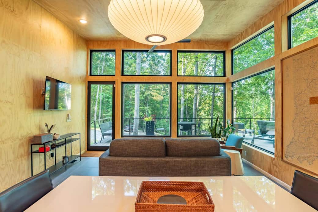 Living room with floor-to-ceiling woodland views, a CB2 white dining table, and a Herman Miller Nelson Bubble light. Modern plywood walls and airy design create the perfect space to relax or gather.
