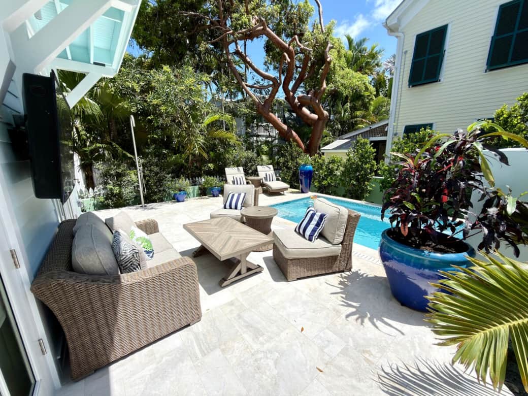 A private outdoor retreat centered around a saltwater pool containing a waterfall feature surrounded by an outdoor shower, lounge chairs, tables, sofa, 2 sitting chairs, and a TV. Designed for relaxing, gathering, and enjoying the Key West sunshine.