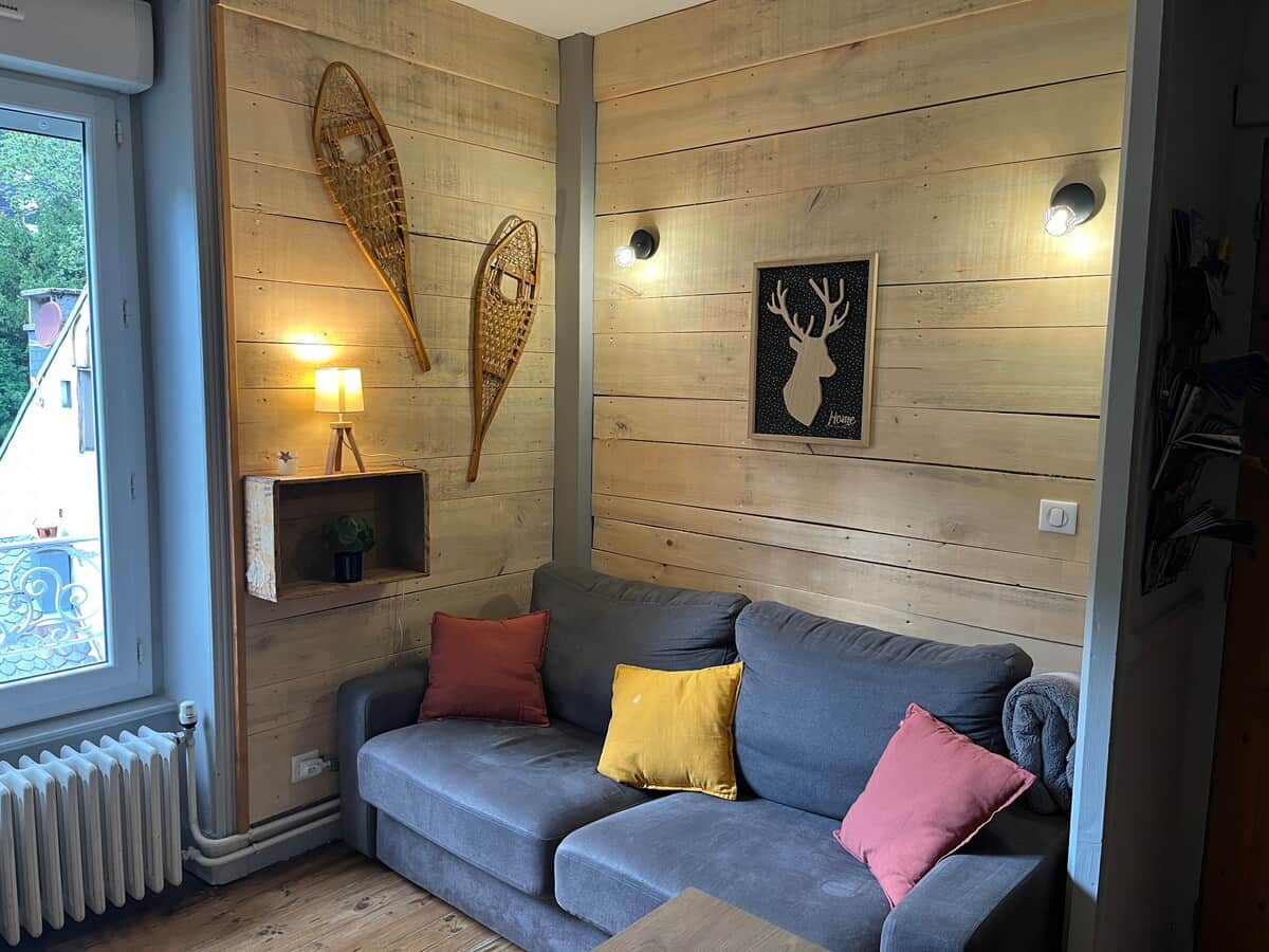 Salon chaleureux au Mont-Dore, ambiance chalet avec murs en bois clair, déco montagnarde (raquettes, ski), canapé gris cosy et lumière douce. Parfait pour un séjour cocooning à la montagne.