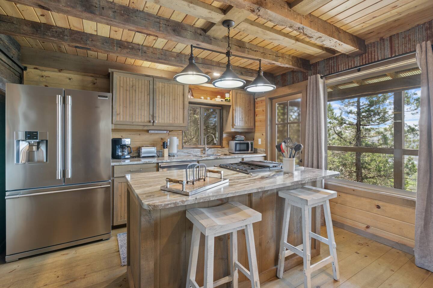 A wide view of a kitchen with a large island, stainless steel appliances, and a window showing trees outside.