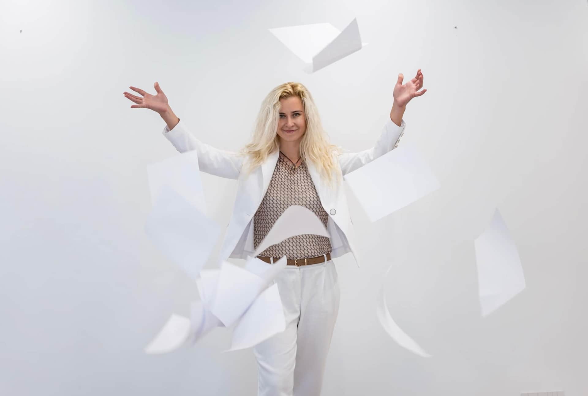 Anastasia Duke laughing as she throws a stack of paper notes into the air inside the RENTAL12 office in Olbia — a glimpse of working method behind the seventeen paintings