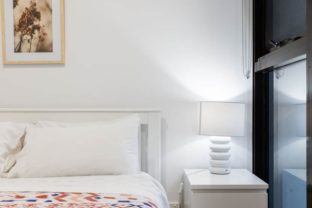 The guest bedroom is a peaceful sanctuary, featuring a comfortable bed with floral linens and a warm, welcoming lamp. The framed artwork adds a personal touch, and the window ensures the room is filled with natural light. 