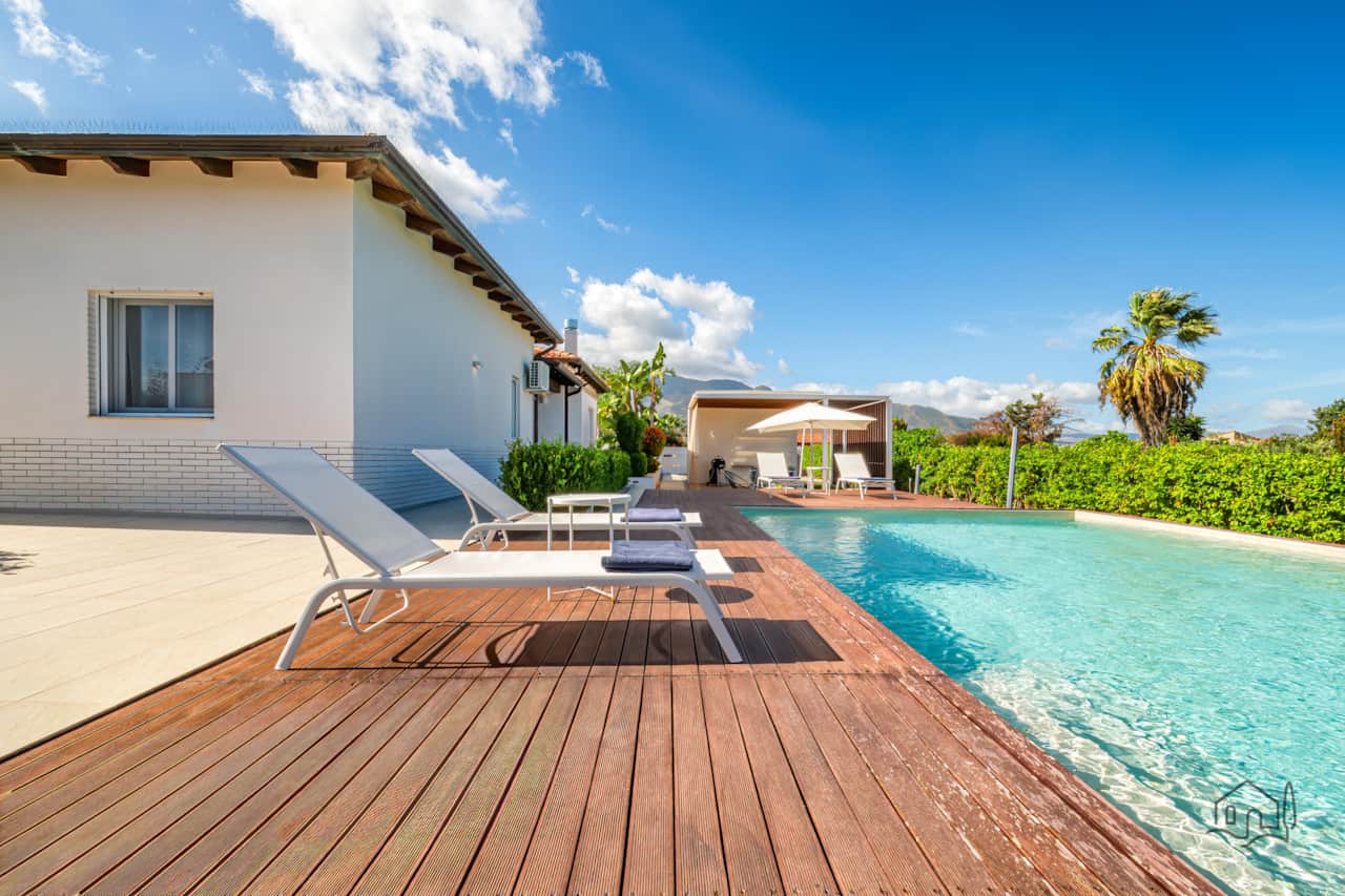 Sunlit pool deck with loungers, with the villa and a shaded dining area in the background.