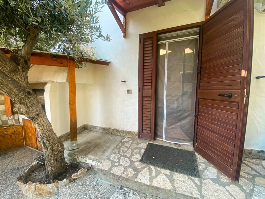 Ingresso rustico ombreggiato da un ulivo, con zanzariera e pavimento in pietra.