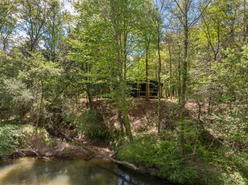 The black cabin feels like it was meant to disappear into the woods—bold in design, but quiet in presence. Perched above the creek and tucked among the trees, it blends into the landscape while still offering a commanding view of the water below.