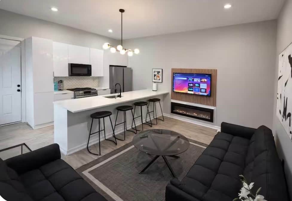 Stylish  living space featuring two futon sofa beds, a flat-screen TV, and a modern electric fireplace. The large island bar offers generous seating and flows into the kitchen, creating a comfortable space to relax, dine, or entertain.