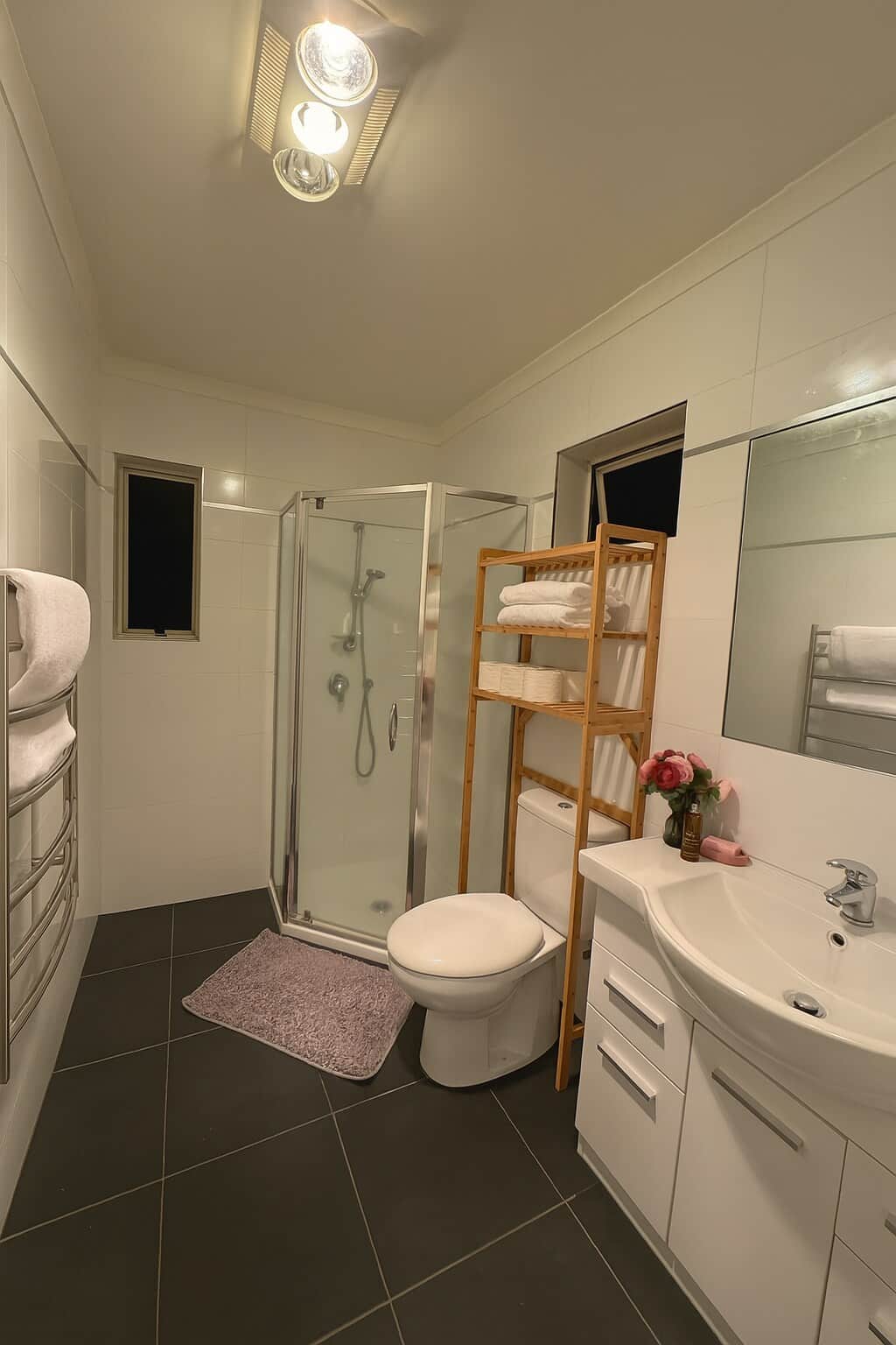 This modern ensuite offers convenience and privacy, featuring a glass shower, sleek vanity, and toilet with open shelving stocked with fresh towels and essentials. A heated towel rail adds a touch of comfort.