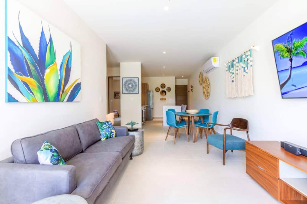 Bright and modern living room with vibrant décor, comfy seating, smart TV, Bose speaker, air conditioning, and a ceiling fan. The open layout connects to the dining area and kitchen, creating a relaxing space after a day at the beach.
