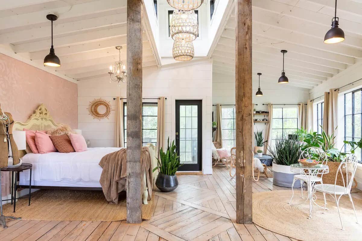 Beautiful open-concept bedroom filled with natural light, featuring elegant chandeliers, rustic wood beams, and lush greenery. Soft pink accents and boho-chic decor create a serene, romantic atmosphere.
