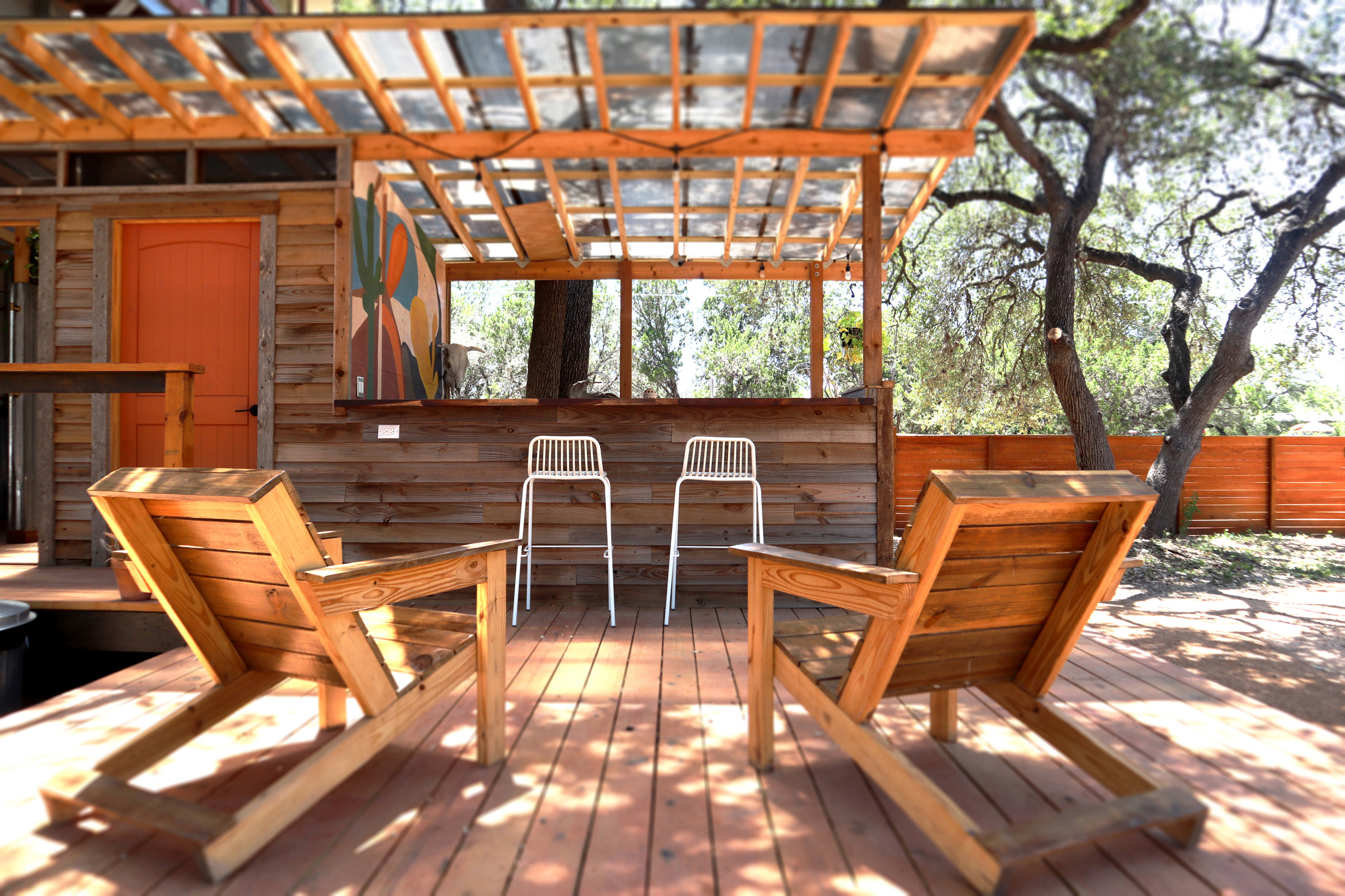 A wooden deck with two wooden chairs and a bar area with two white stools under a pergola.