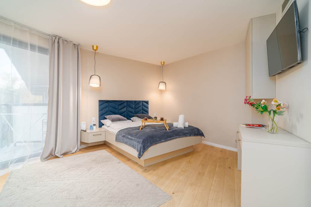 Przytulna, jasna sypialnia z dużym łóżkiem i granatowym zagłówkiem, sofą oraz wyjściem na balkon.