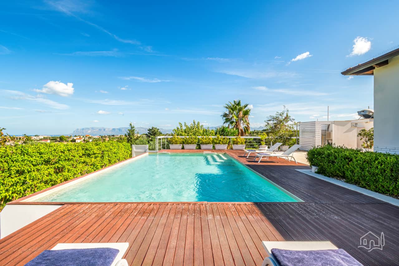 Panoramic pool deck with sun loungers, lush greenery, and distant sea views on the horizon.