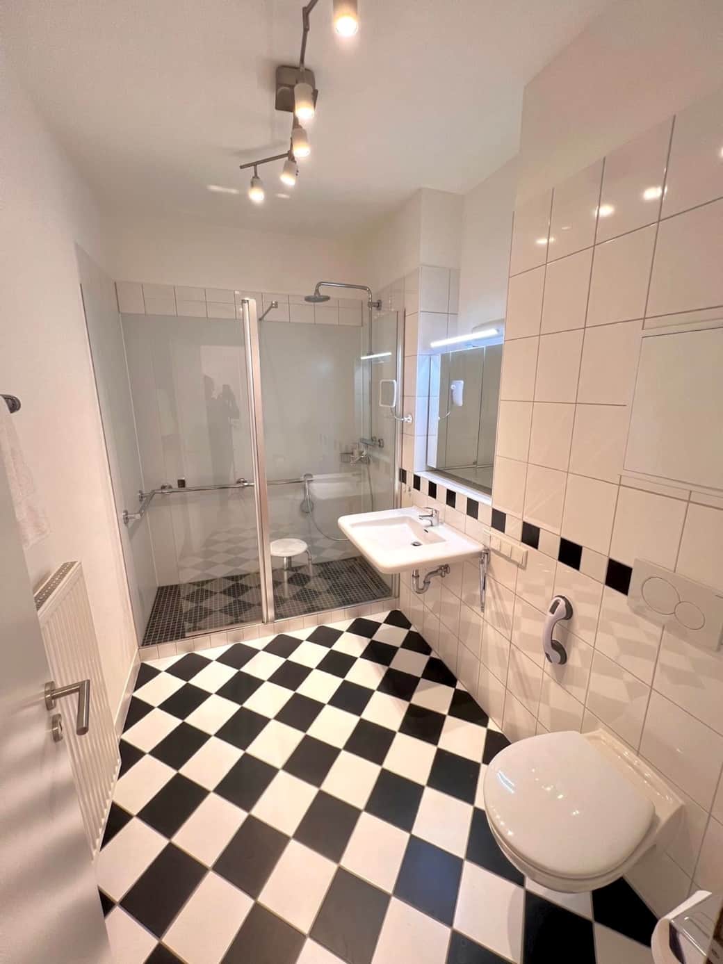 Das klassisch eingerichtete Badezimmer verfügt über eine großzügige Dusche, frischen Handtüchern, ein unterfahrbares Waschbecken mit dreitürigen Spiegelschrank sowie eine leicht erhöhte Toilette mit verstellbaren Haltegriffen. 