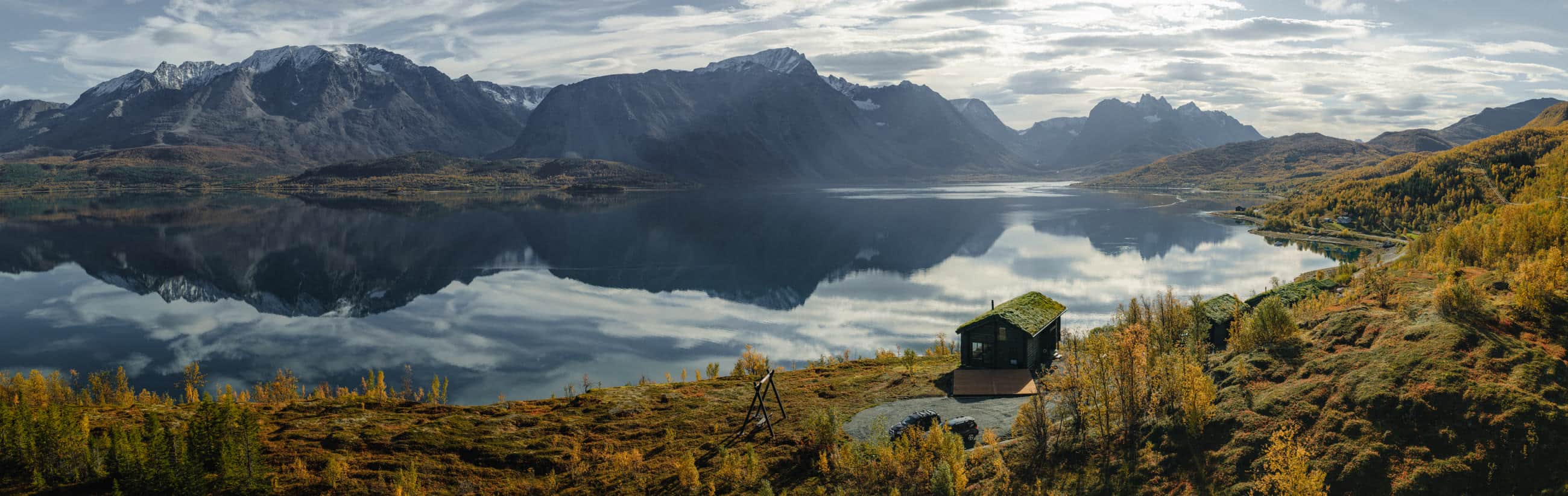 Blockhaus mit Gründach am Hang, darunter der spiegelglatte Fjord, gegenüber die Lyngen-Alpen