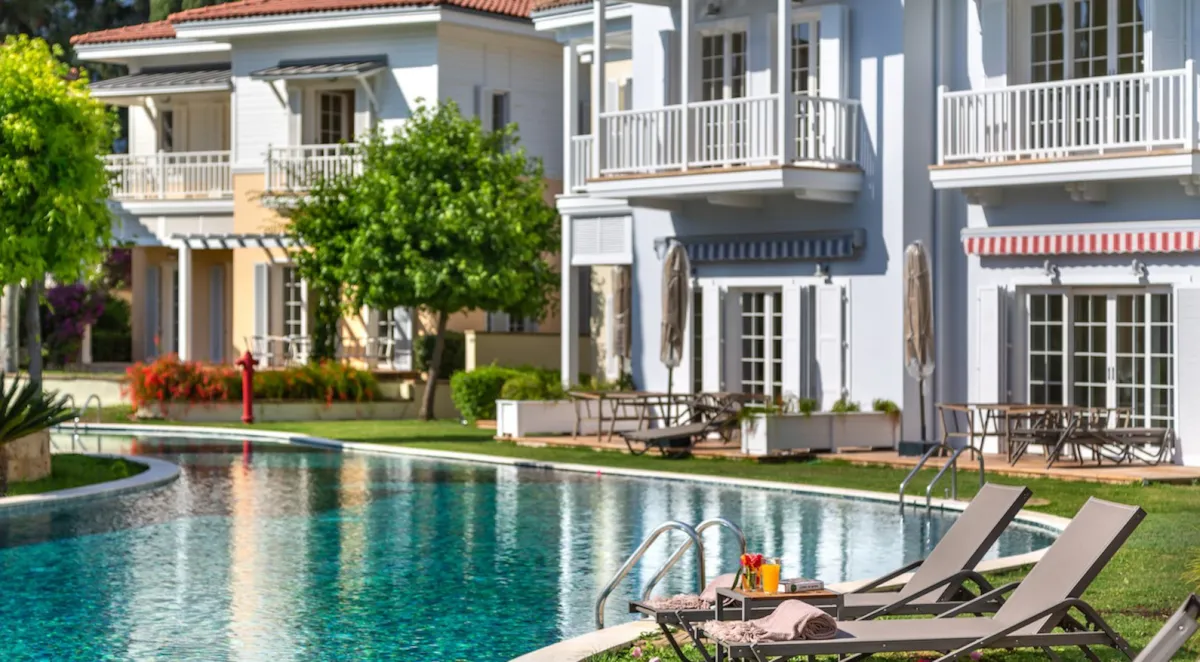 Swimming pool area with sun loungers and a table, surrounded by greenery and residential buildings in Göcek.