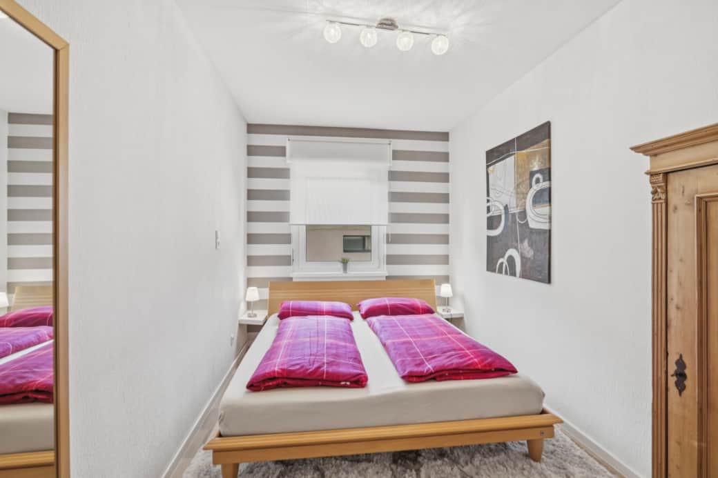 Doppelbett mit pinken Kissen auf Holzrahmen, grau-weiß gestreifte Wandtapete, dezente Deckenbeleucht