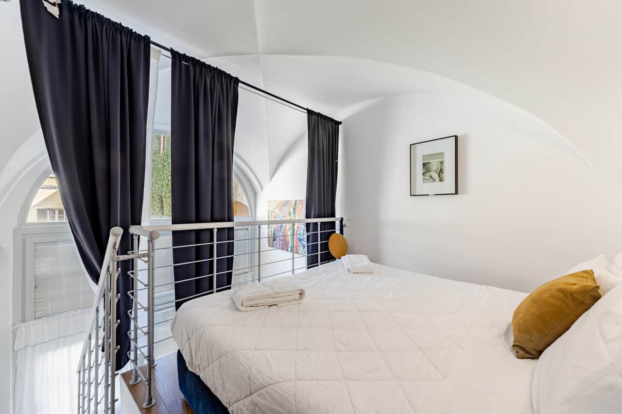 Schlafzimmer mit weißem Bett, dunklen Vorhängen und bodentiefem Fenster