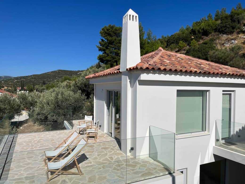 The Studio Apartment :A sun-drenched terrace unfolds around a whitewashed house with terracotta roof tiles. Lounge chairs overlook olive trees and forested hills, offering a peaceful retreat where sky, landscape & architecture meet holiday serenity.