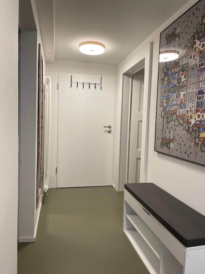 Couloir avec un meuble à chaussures, un grand miroir et un porte manteau.
Vue depuis la porte d'entrée avec à droite, les chambres et à gauche la cuisine et la salle de bain.