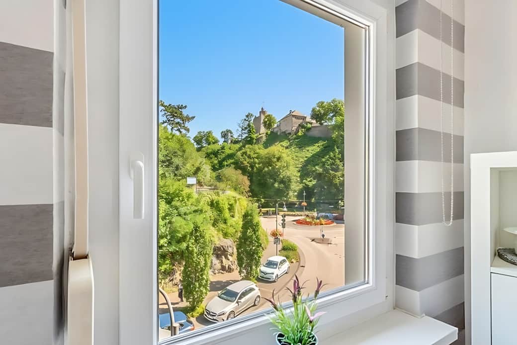 Blick durch ein weißes Fenster auf die Burg Saarburg, grüne Bäume, Kreisverkehr und blauen Himmel.
