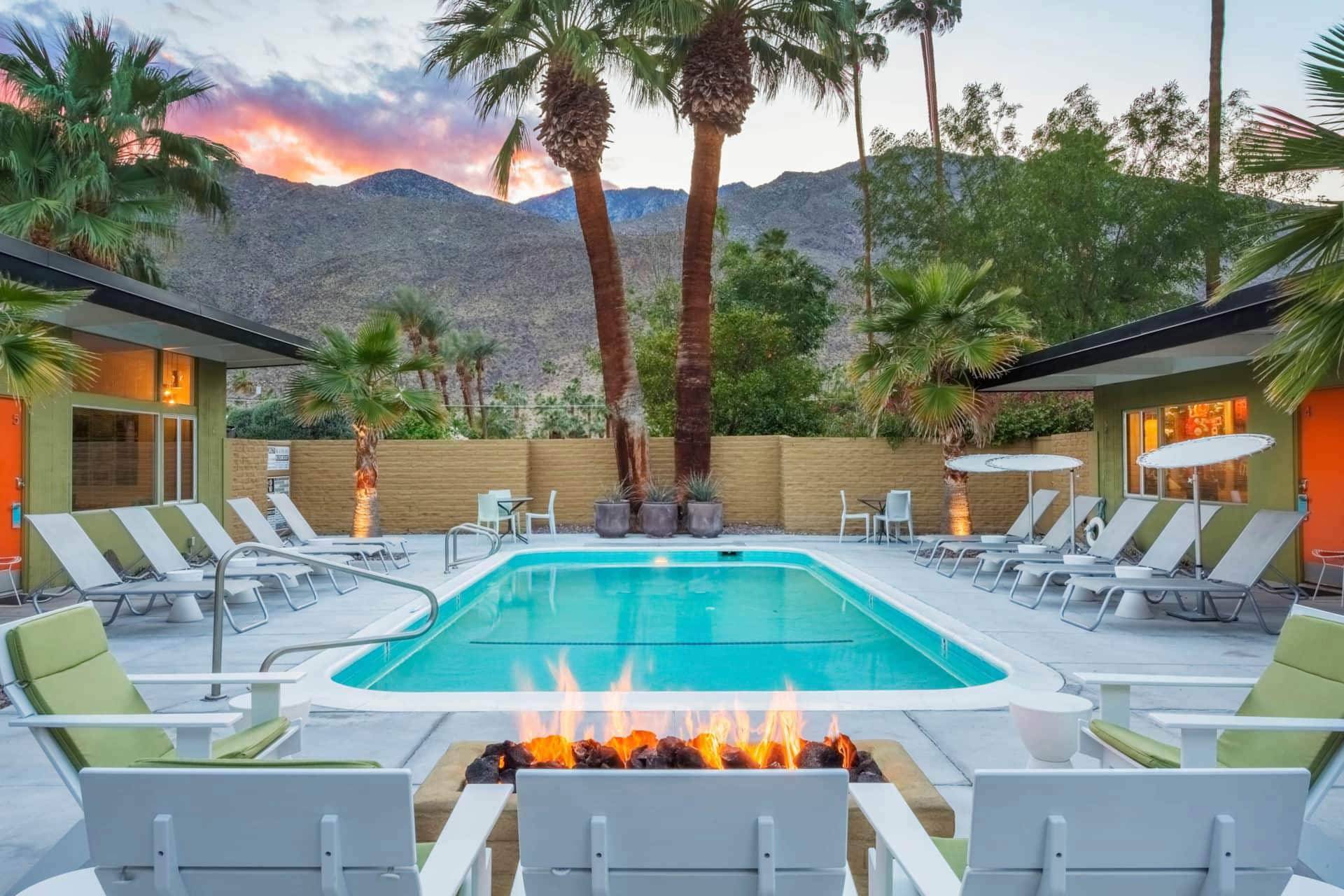 A pool area with a fire pit, lounge chairs, and palm trees in front of a mountain view at sunset.