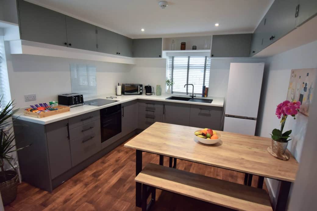 Modern, fully equipped kitchen with sleek grey cabinets, wood flooring, and a large dining bench. Includes oven, microwave, fridge-freezer, toaster, kettle and coffee machine. Bright, clean, and perfect for preparing meals or relaxing with a brew.