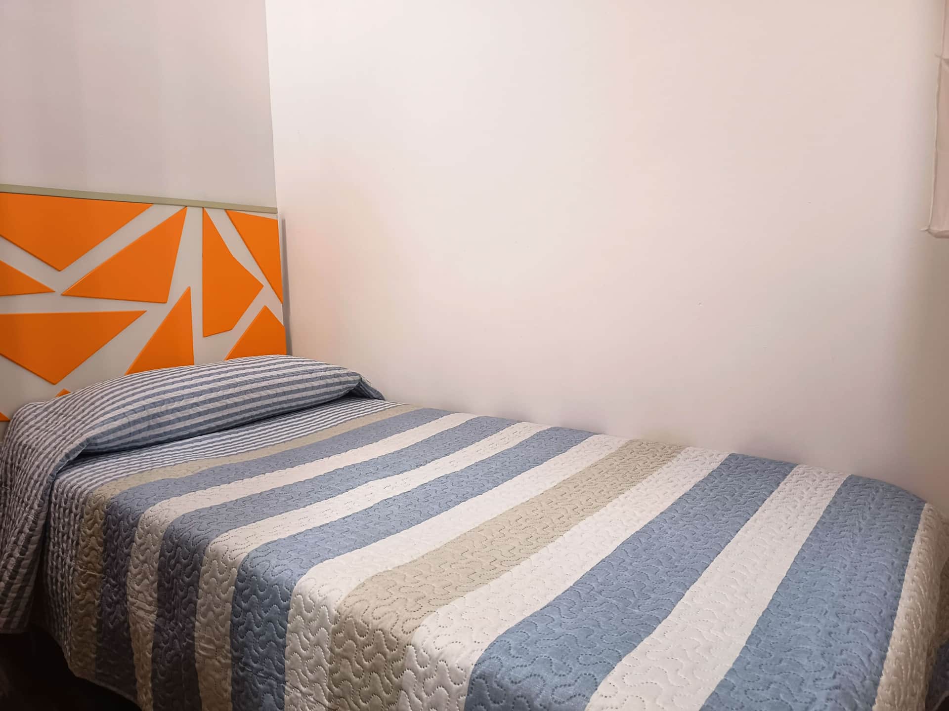 Habitación individual con cama de estilo moderno, cabecero geométrico en tonos naranjas y colcha a rayas azules y beige