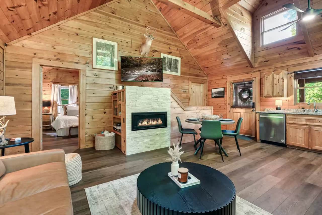 Warm, open-concept cabin interior with vaulted wood ceilings, a cozy fireplace, and full kitchen