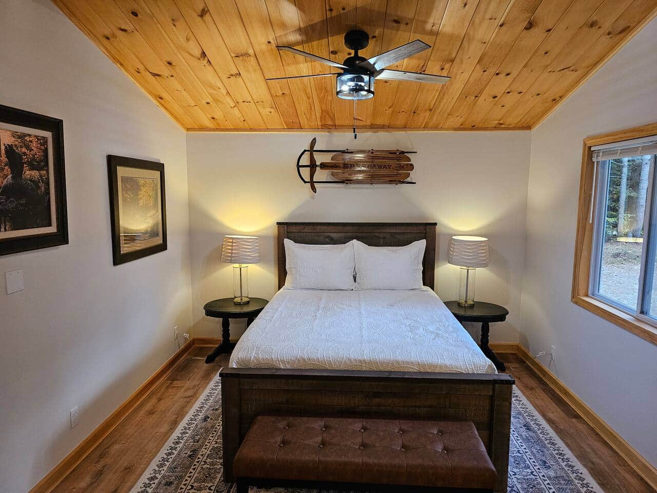 Master retreat with queen bed under a T&G ceiling with fan and lights. Matching glass-base lamps on side tables, leather bench, and large rug add comfort. Vintage toboggan and wildlife art accent the room; big windows with louvered shades.