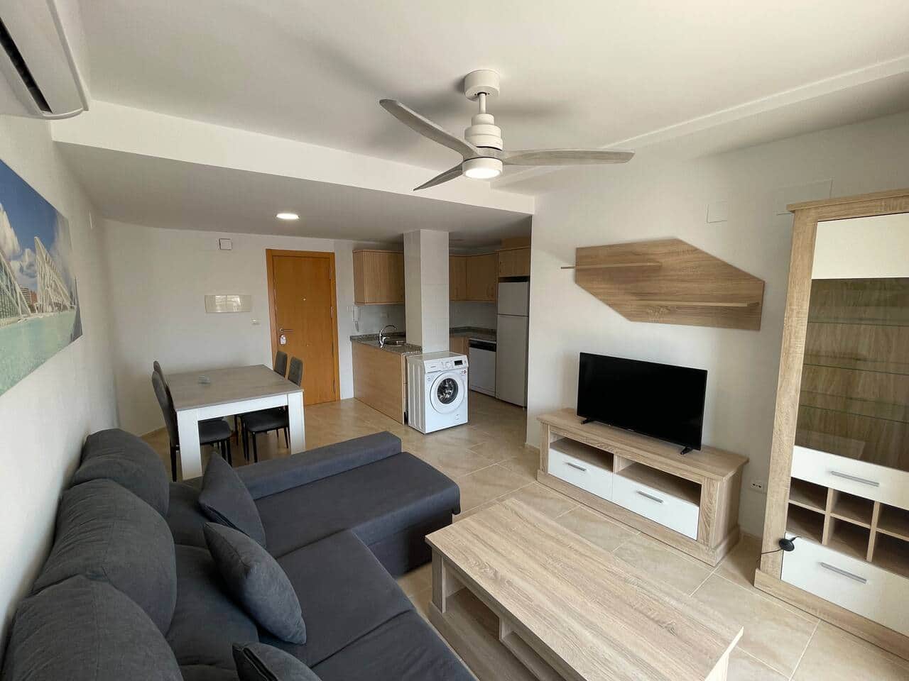Salón con sofá rinconera, TV y cocina abierta con lavadora