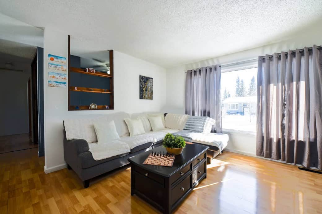 A bright living room with large windows, a cozy sectional sofa with soft throws, and a stylish coffee table with a chess set. Enjoy the natural light, wooden floors, and a peaceful space to relax.