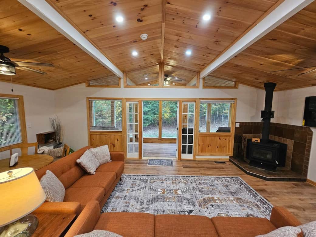 Spacious living room with wood stove, vaulted tongue-and-groove ceiling, and forest views. Relax on mid-century sofas by large windows and enjoy warm natural light and cozy modern-rustic style.