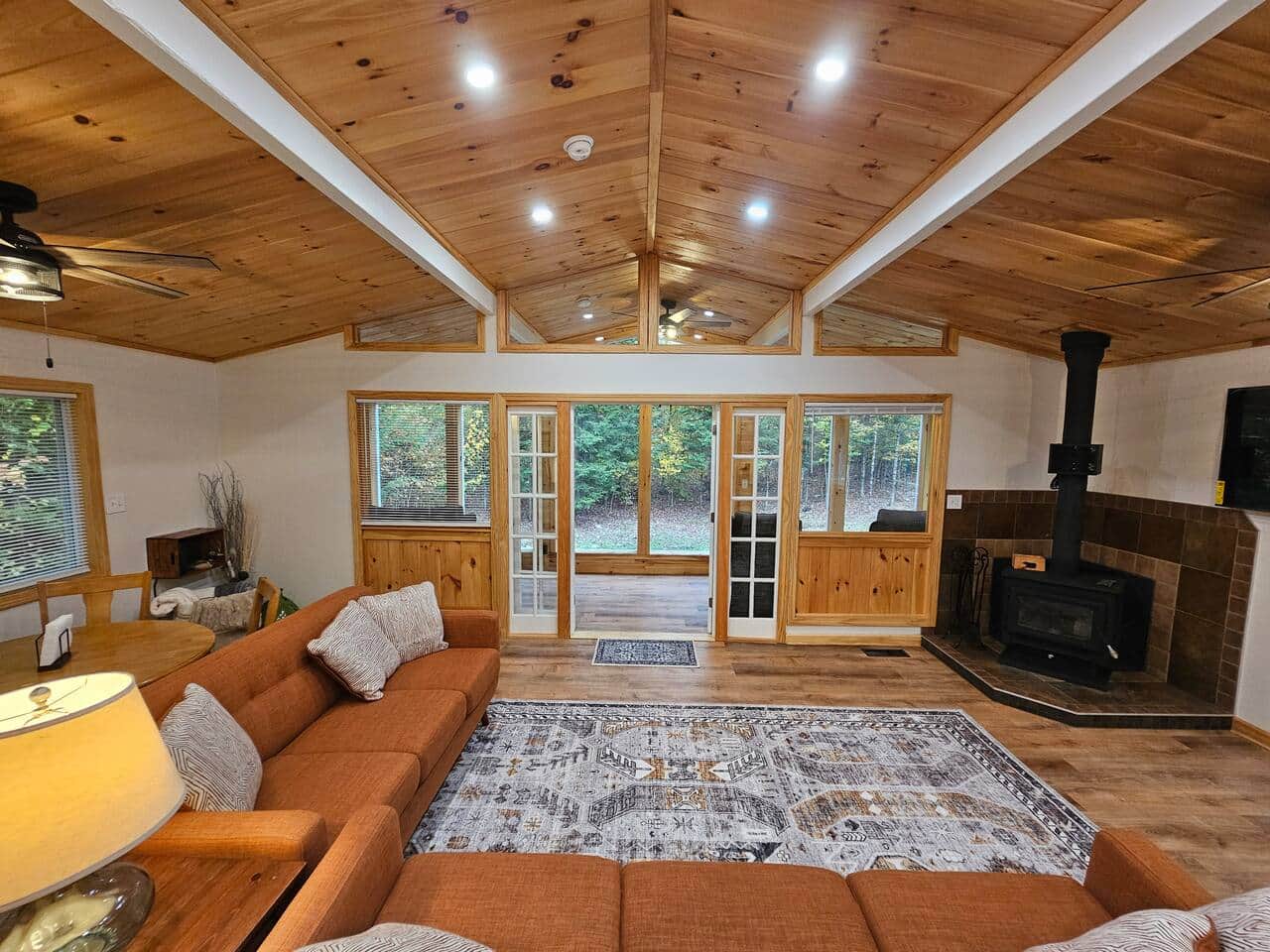 Spacious living room with wood stove, vaulted tongue-and-groove ceiling, and forest views. Relax on mid-century sofas by large windows and enjoy warm natural light and cozy modern-rustic style.