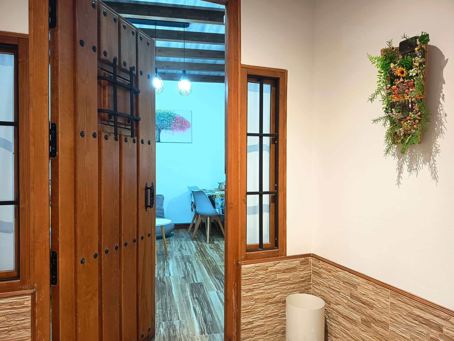 Puerta de madera rústica abierta que da paso a un comedor con mesa y sillas, decoración floral en la pared y suelo de madera