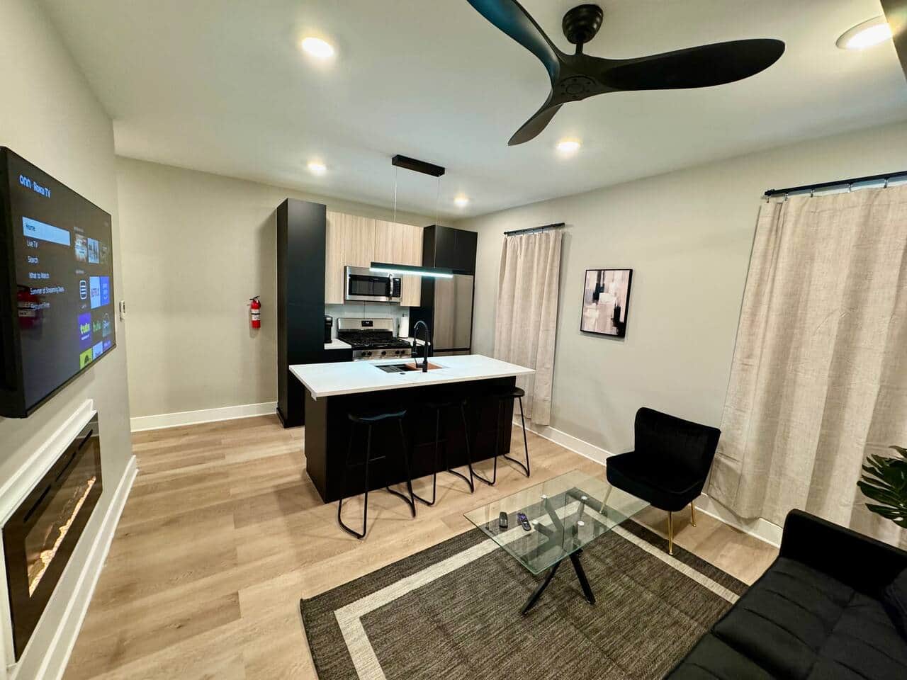 Modern kitchen featuring brand-new appliances, including a breakfast bar, dishwasher, microwave, and refrigerator. It seamlessly connects to the living room, which is furnished with a sofa bed, ana a large flat-screen TV.
