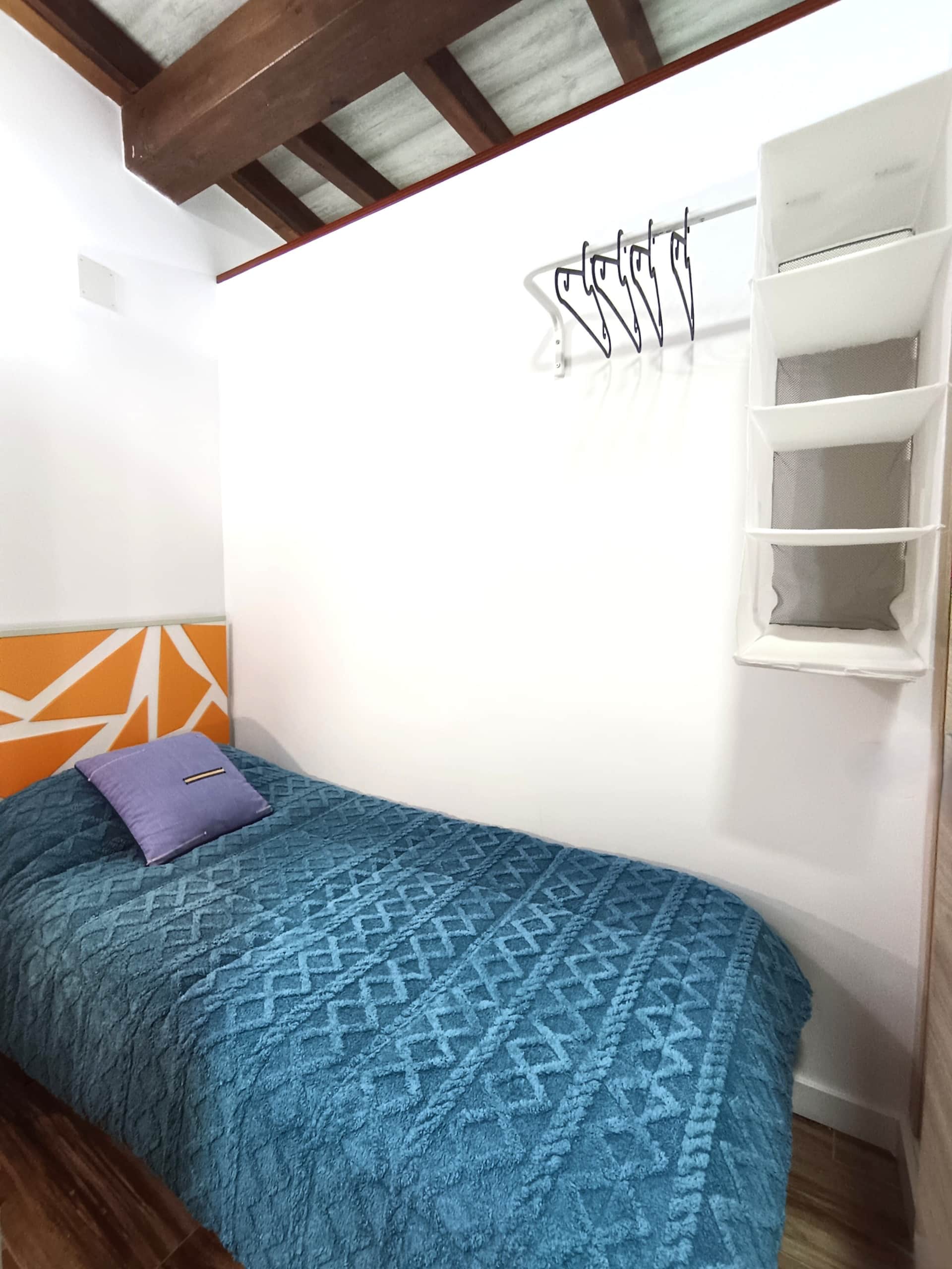 Habitación individual acogedora en casa rural en Polán, cama con colcha azul geométrica y cabecero en tonos naranja y blanco, barra con perchas y organizador